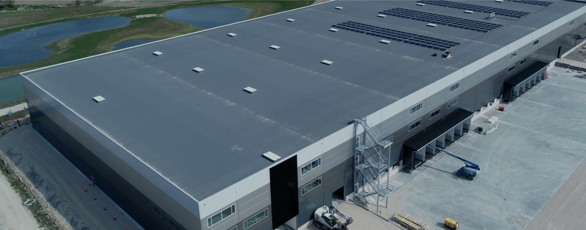 aerial view of a grey and blue warehouse with scaffolding and a series of small lakes behind it